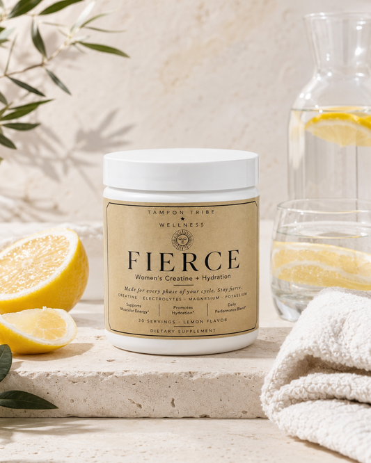 A white plastic jar of Tamron Tribe Wellness Fierce dietary supplement with a beige label, styled on a stone surface with lemon slices, a glass of water, and a towel.