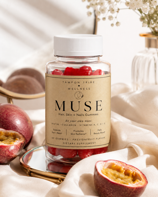A clear bottle of TAMPON TRIBE WELLNESS MUSE Hair, Skin + Nails Gummies with red gummies visible inside, set on a gold tray with a mirror. The scene is styled with passionfruit, draped white fabric, and soft background lighting.